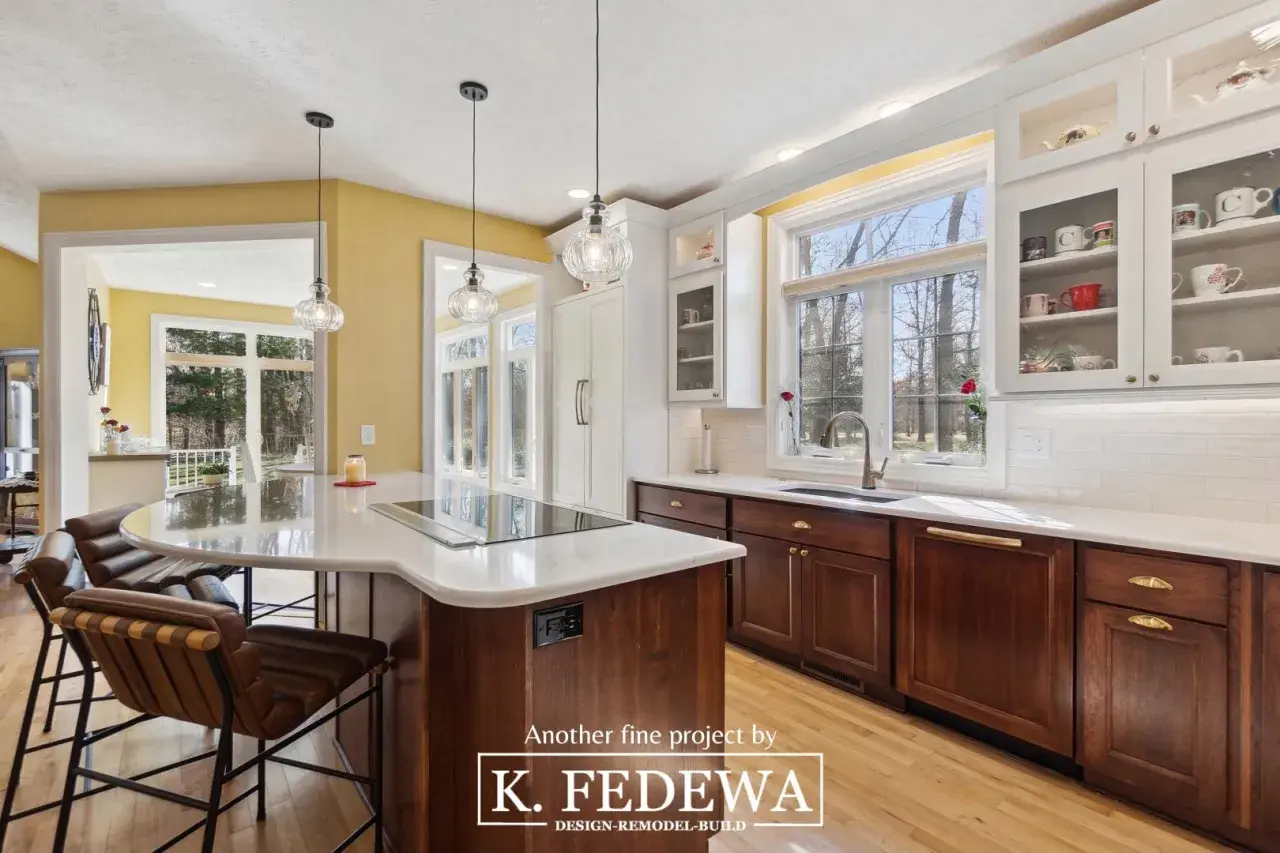 A bright, airy kitchen with natural wood cabinets and a large island. Sunlight streams through large windows, illuminating the warm tones of the wood.