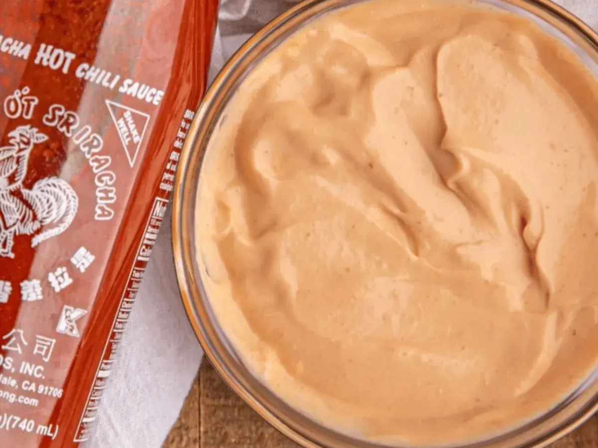 A bowl of creamy, orange spicy mayo recipe sits next to a bottle of Sriracha hot chili sauce.