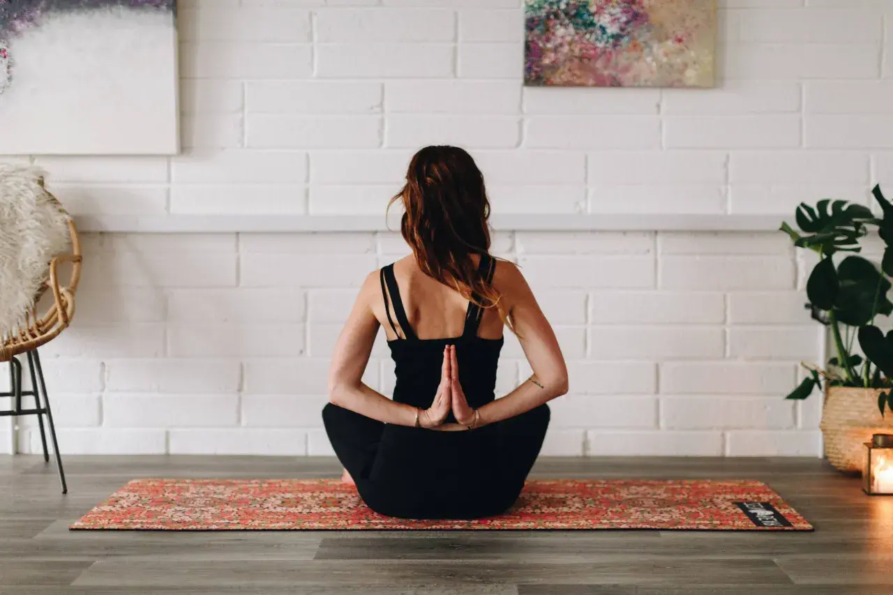 Femme en tenue noire pratiquant le hatha yoga, mains jointes derrière le dos, assise sur un tapis orné.