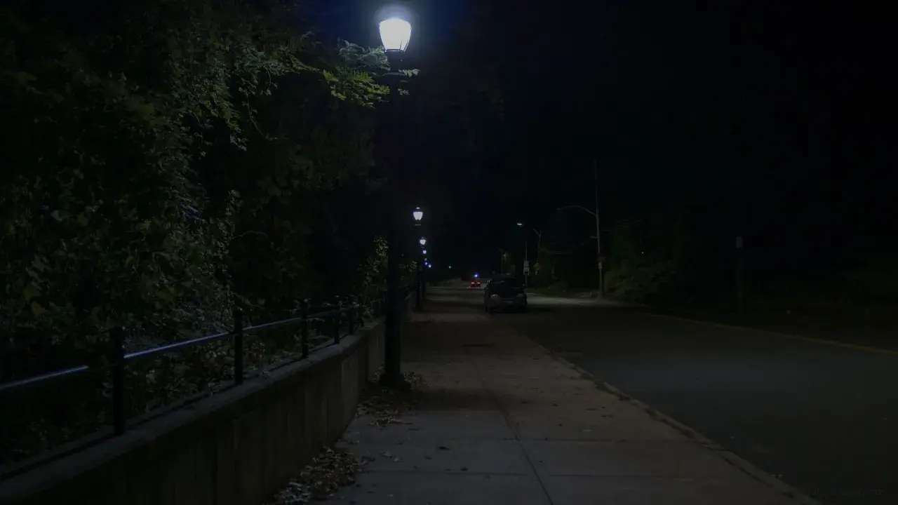 Nocna ulica z latarniami, chodnikiem i samochodami. Ciemność i światło lamp tworzą atmosferę jak z filmu, gdzie nawet kurza ślepota wydaje się możliwa.