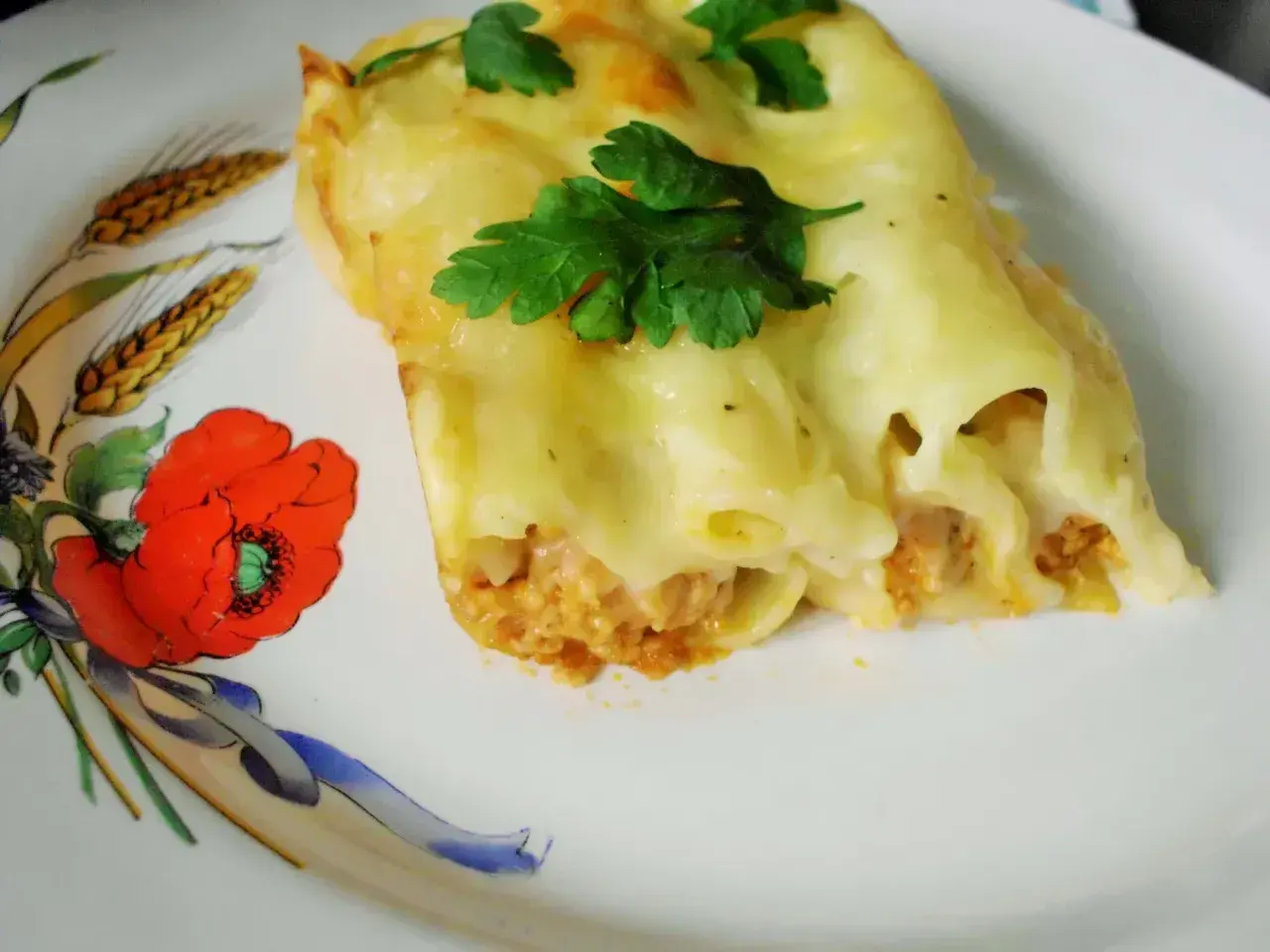 Pyszne cannelloni z mięsem mielonym, zapieczone pod serem i udekorowane natką pietruszki. Danie podane na talerzu z motywem maków i kłosów.