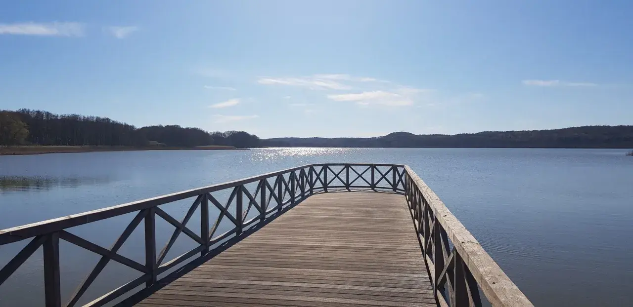 Holzsteg am Schmachter See lädt zum Spaziergang auf dem Rundweg ein. Sonnenschein glitzert auf dem Wasser, bewaldete Ufer im Hintergrund.
