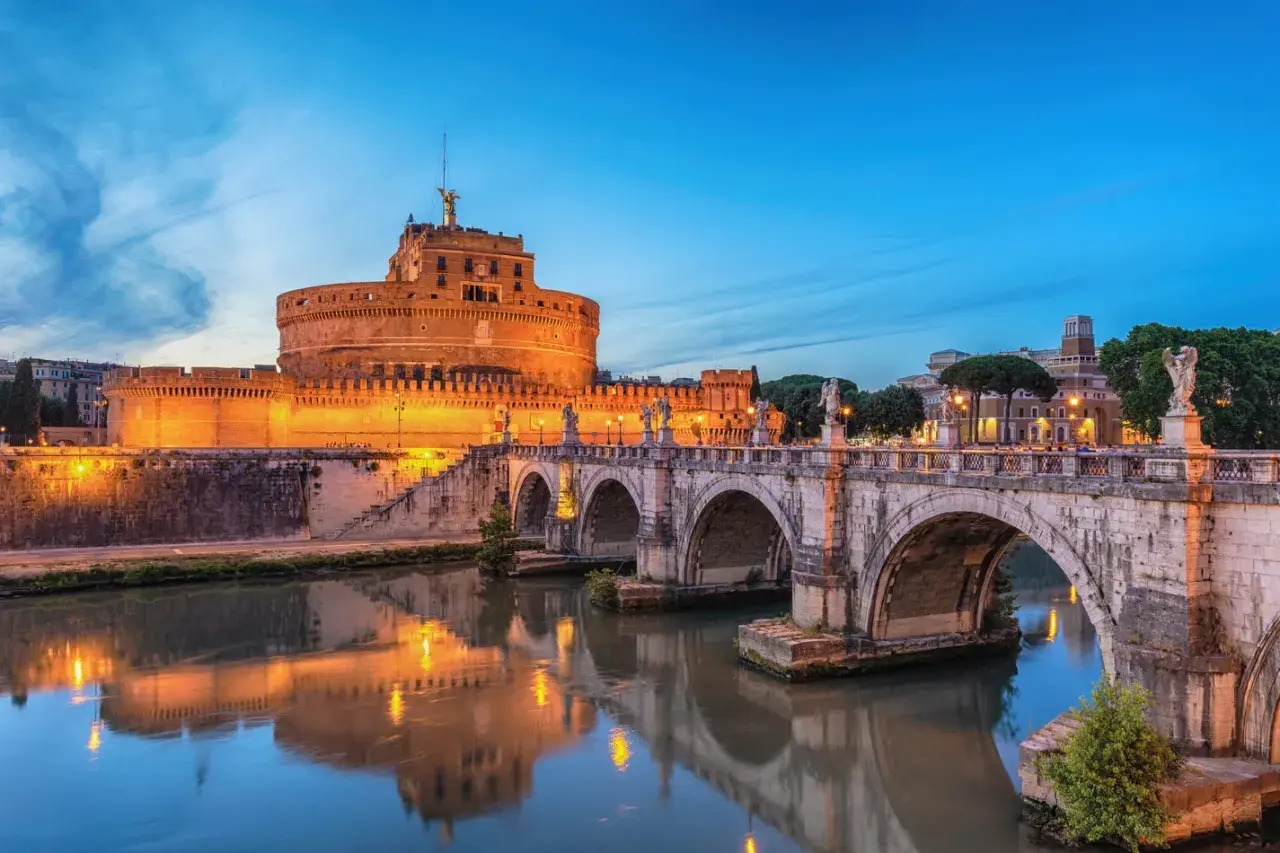 Le Castel Sant'Angelo à Rome, illuminé au crépuscule, se reflète dans le Tibre. Le pont Saint-Ange, orné de statues, mène à cette ancienne forteresse.