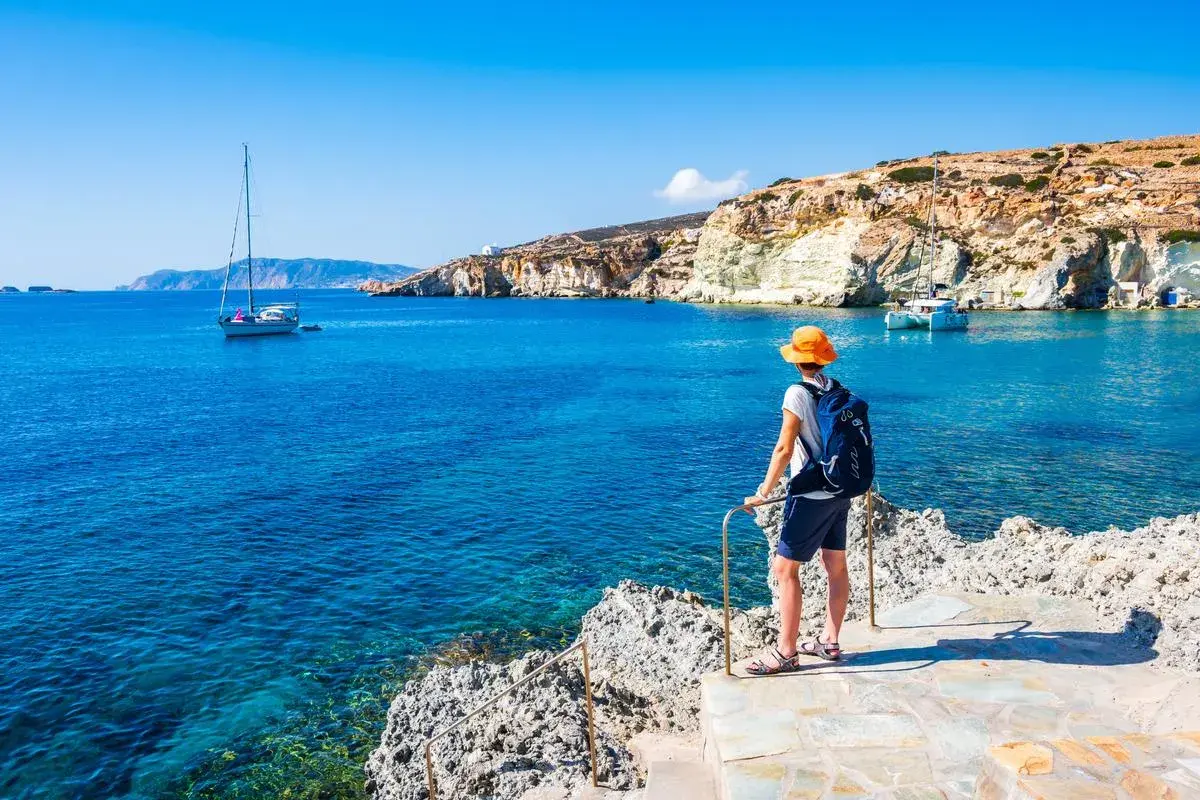 Un voyageur admire la mer Égée bleue et les falaises blanches, un voilier au loin. Le tourisme en Grèce offre des paysages magnifiques.