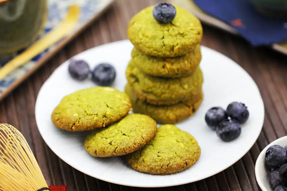 Grüne matcha kekse mit Blaubeeren auf einem Teller, daneben ein Matcha-Schneebesen.