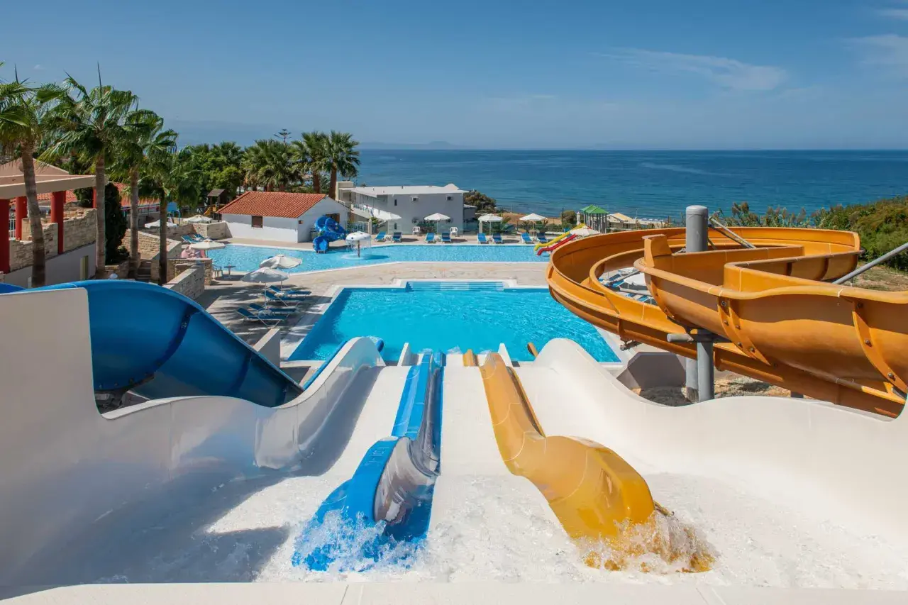Vattenland på Kreta med färgglada rutschkanor som leder ner i en stor pool. Havet glittrar i bakgrunden.