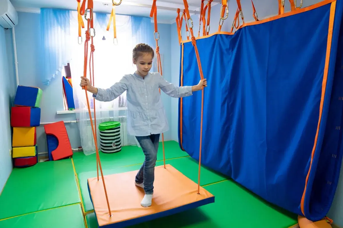 Dziewczynka na huśtawce w sali do terapii sensorycznej. Terapia sensoryczna na czym polega? Na stymulacji zmysłów poprzez ruch i zabawę.
