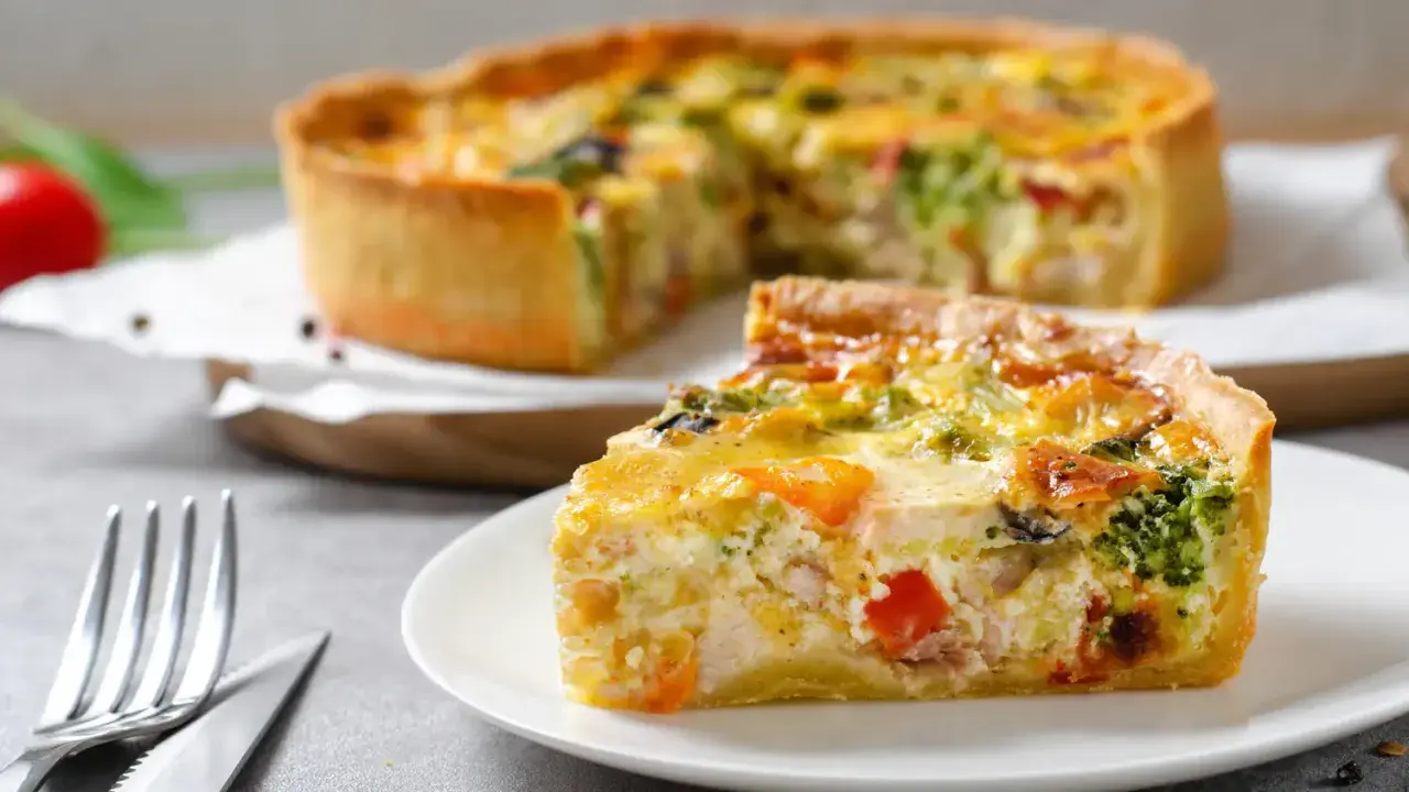 Une part de quiche savoureuse avec une pâte à tarte maison dorée, garnie de légumes et de poulet. Le reste de la tarte est flou en arrière-plan.