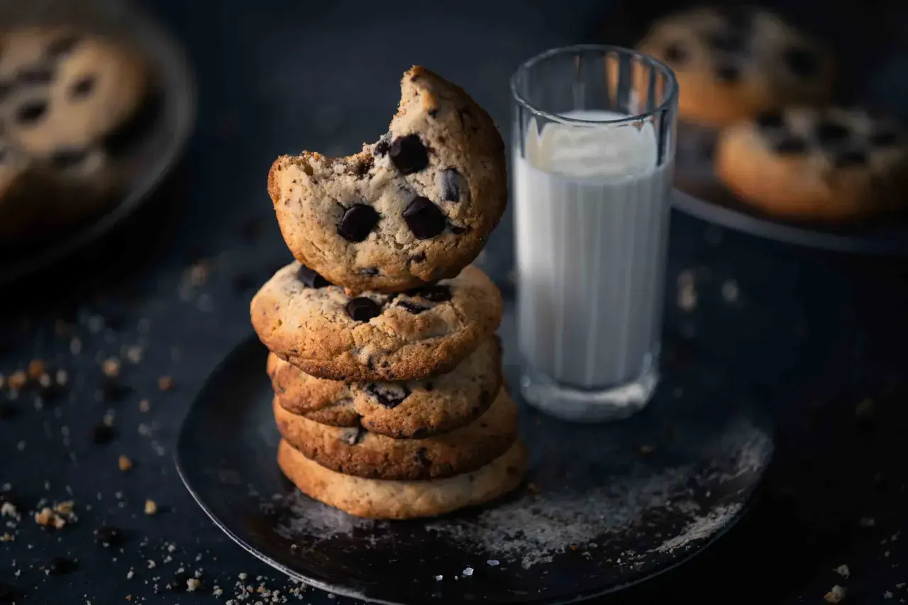 Ein Stapel Schokoladenkekse, eines davon angeknabbert, neben einem Glas Milch. Perfekt für ein einfaches Cookies Rezept.