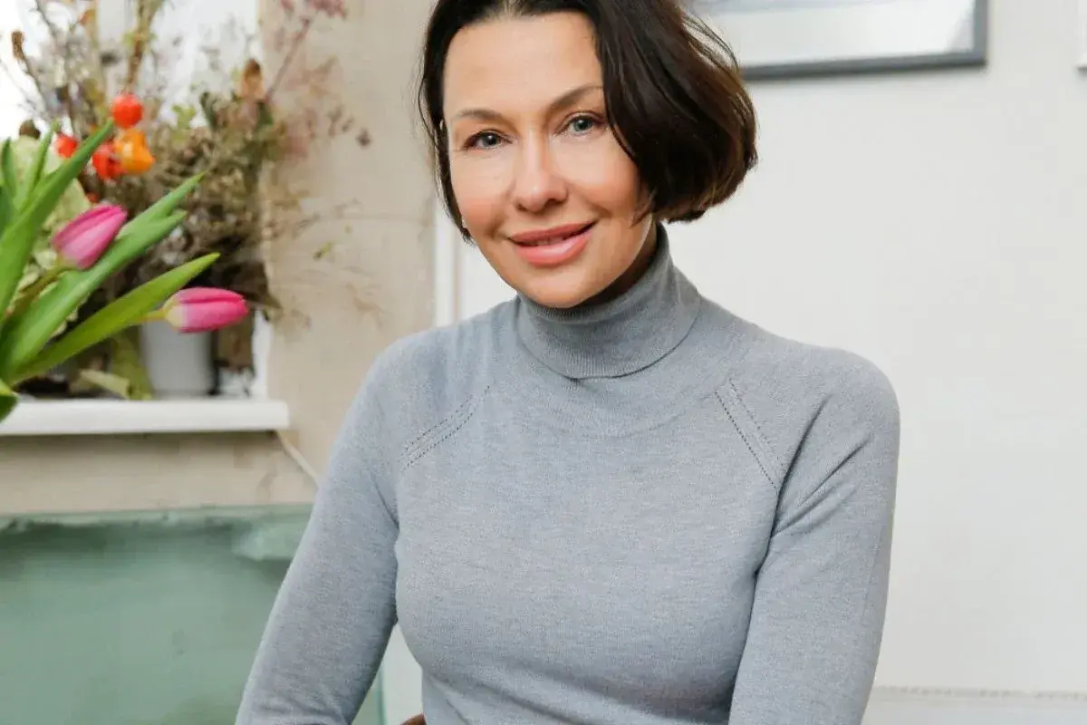 Uśmiechnięta Anna Popek w szarym golfie, obok bukiet różowych tulipanów. Wiek nie jest widoczny, ale jej uroda zachwyca.