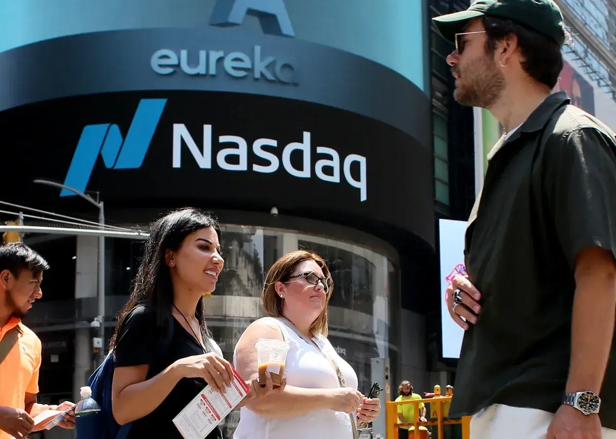 Ludzie przed budynkiem Nasdaq, gdzie handluje się akcjami. Słońce świeci, tworząc cień.