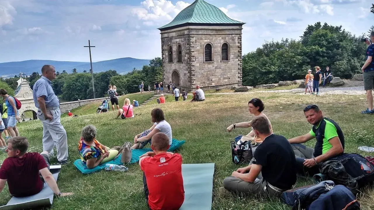 Ludzie odpoczywają na trawie pod kamienną wieżą i krzyżem. Trasa na Święty Krzyż przyciąga turystów.