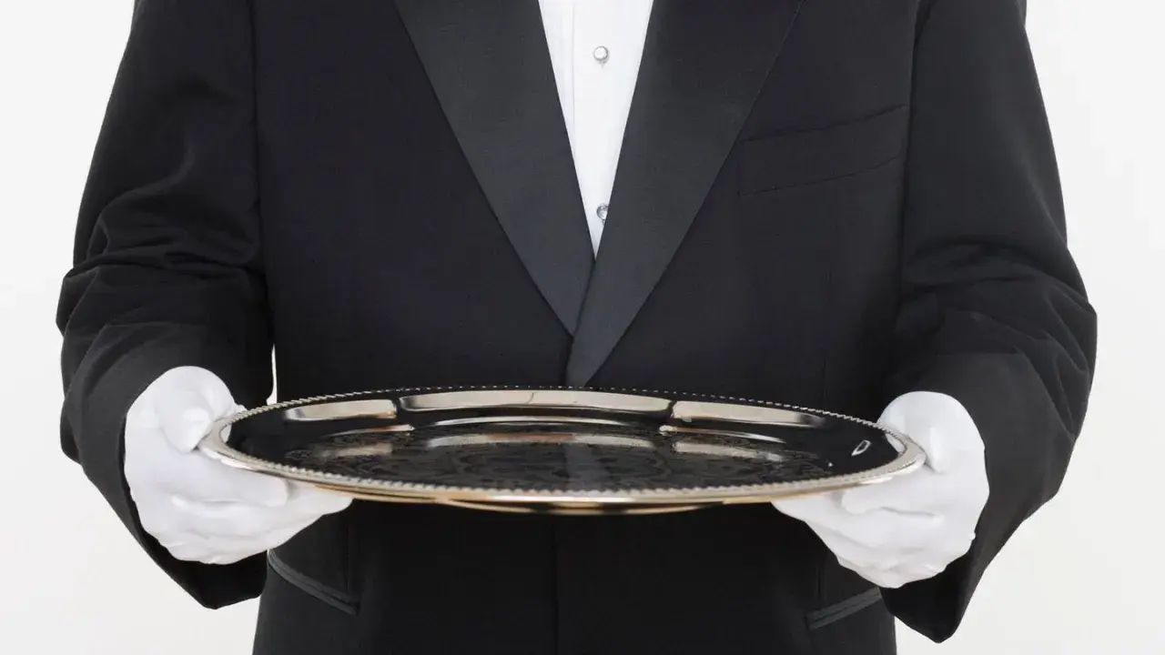 Ein Butler in schwarzem Frack mit weißer Weste und Handschuhen hält ein Silbertablett. Der Butler gehalt ist ein wichtiger Faktor für den Service.