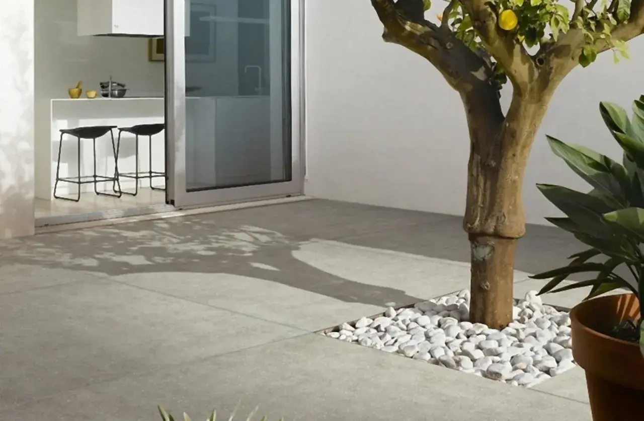 Un moderno patio con diversi tipi di pavimentazione esterna: lastre in gres grigio e un'area con ciottoli bianchi attorno a un albero.