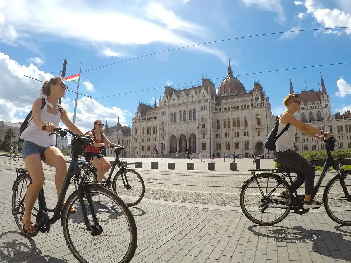 Friends enjoying Budapest bike tours, cycling past the Parliament building under a bright, cloudy sky.