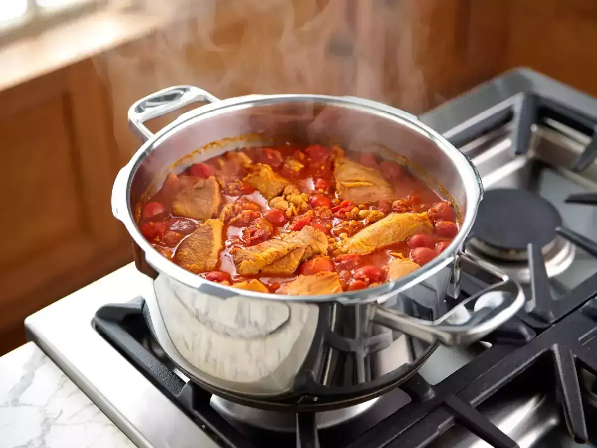 Un délicieux saute de porc mijote dans une cocotte en acier inoxydable sur une cuisinière à gaz, dégageant une vapeur appétissante.