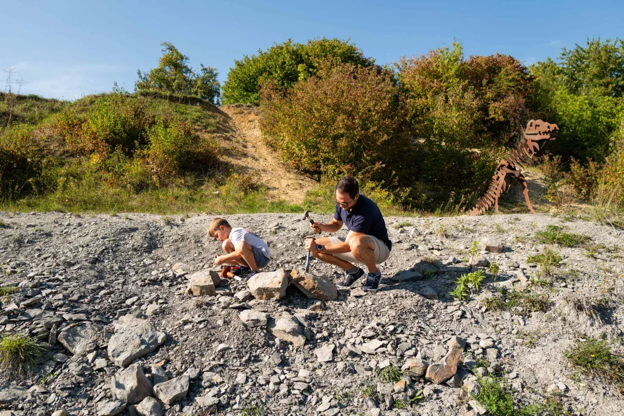 Ein Vater und sein Sohn graben nach Fossilien. Im Hintergrund steht ein Skelett eines Dinosauriers.