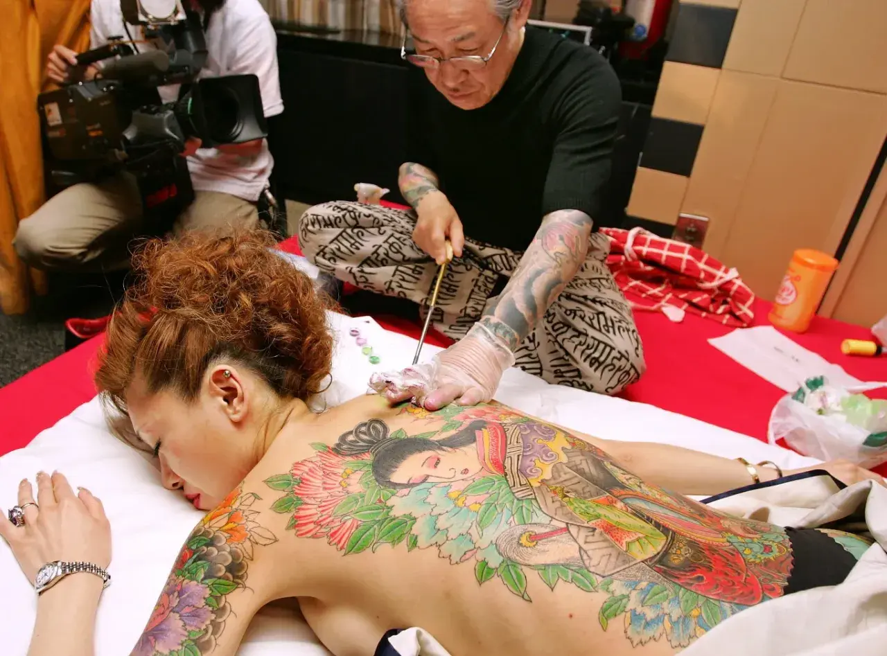 Mistrz tatuażu tworzy tradycyjne tatuaże w Japonii na plecach kobiety. W tle operator kamery dokumentuje proces.