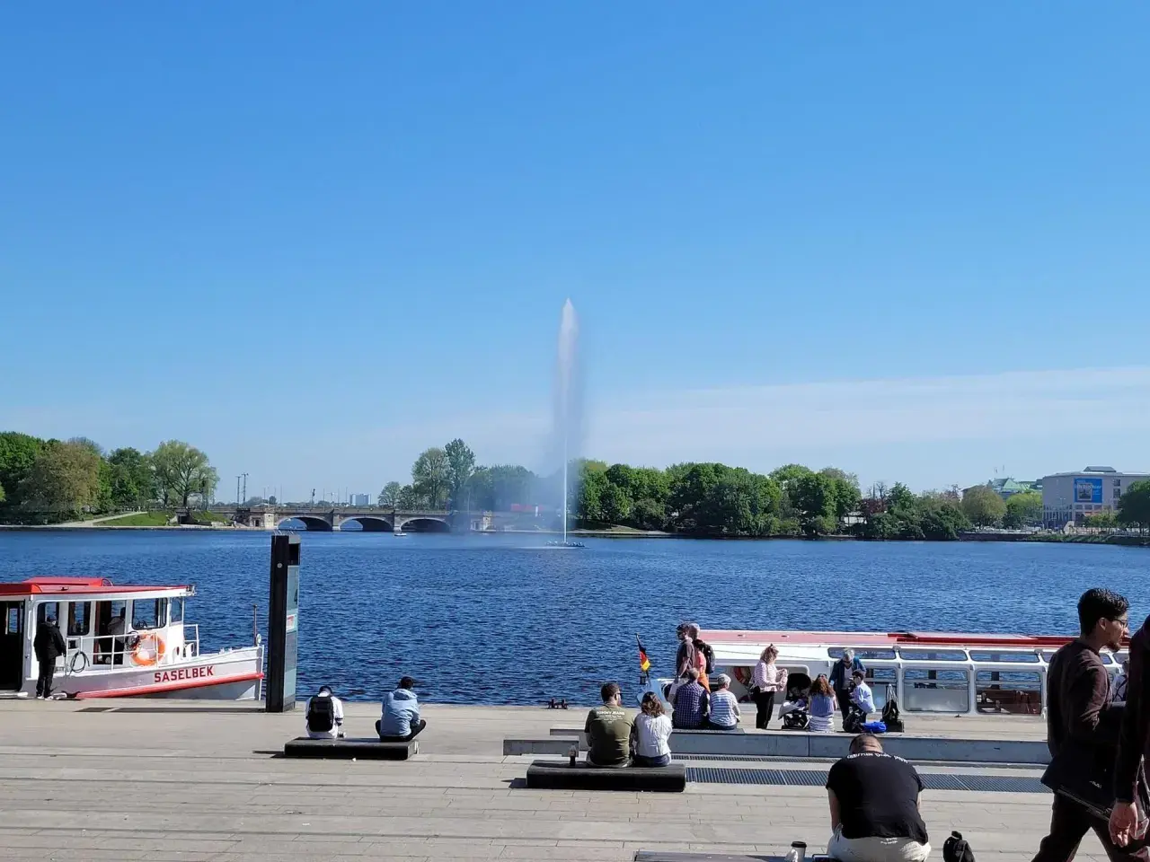 Ein sonniger Tag am Alstersee Hamburg. Menschen entspannen sich am Ufer, während die berühmte Fontäne in die Höhe schießt.
