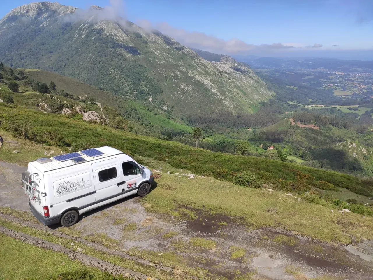 Furgoneta blanca con paneles solares aparcada en una ladera verde, lista para viajar en furgoneta y explorar las montañas.