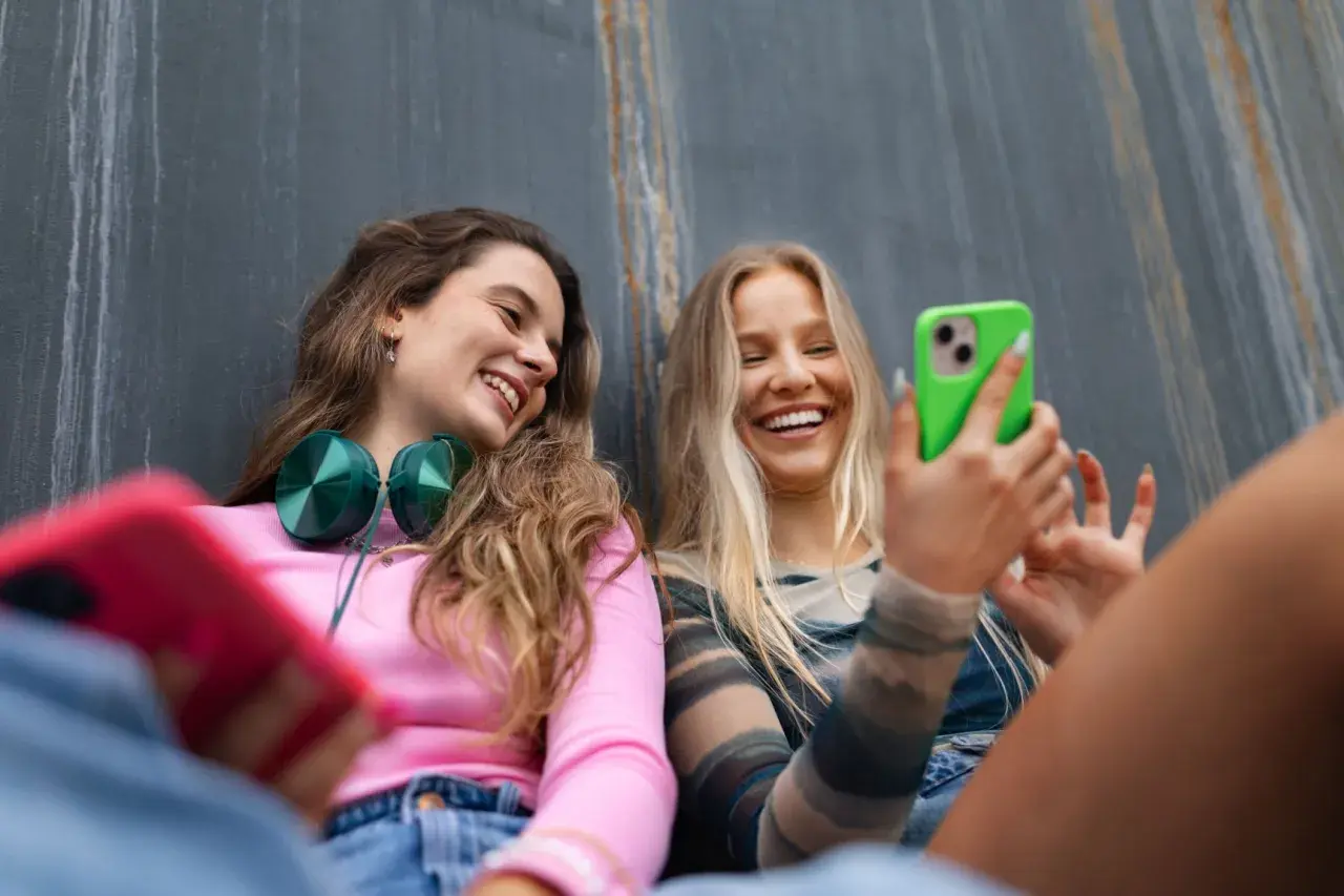 Dwie uśmiechnięte dziewczyny rozmawiają przez **Facetime** na telefonach, jedna z nich trzyma zielony smartfon.