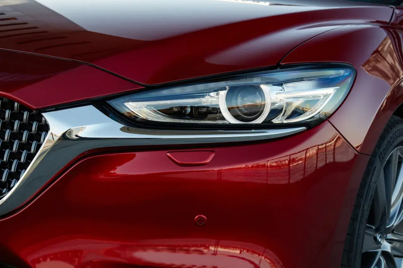 Błyszczący, czerwony lakier Mazda 6 Skyactiv odbija otoczenie. Widoczny fragment reflektora i chromowana atrapa.