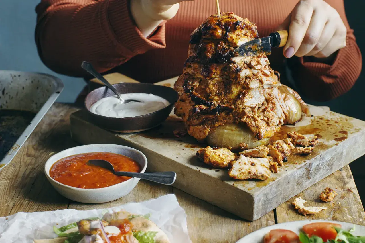 En person skär av en bit från en saftig kyckling shawarma som roterar på ett spett. Tillbehör som vitlökssås och en röd sås finns bredvid.