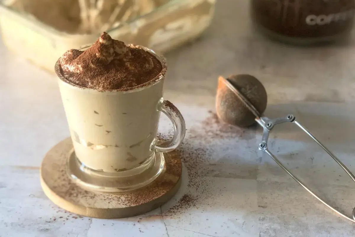 Deliziosa crema al caffè in una tazza di vetro, spolverata di cacao. Accanto, un colino per dolci e un barattolo di caffè.