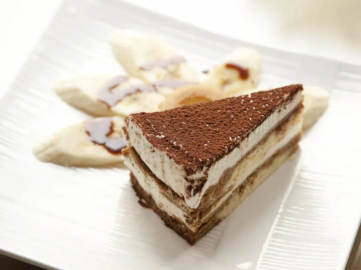 Tranche de tiramisu banane, saupoudrée de cacao, accompagnée de rondelles de banane nappées de sauce. Un délice !