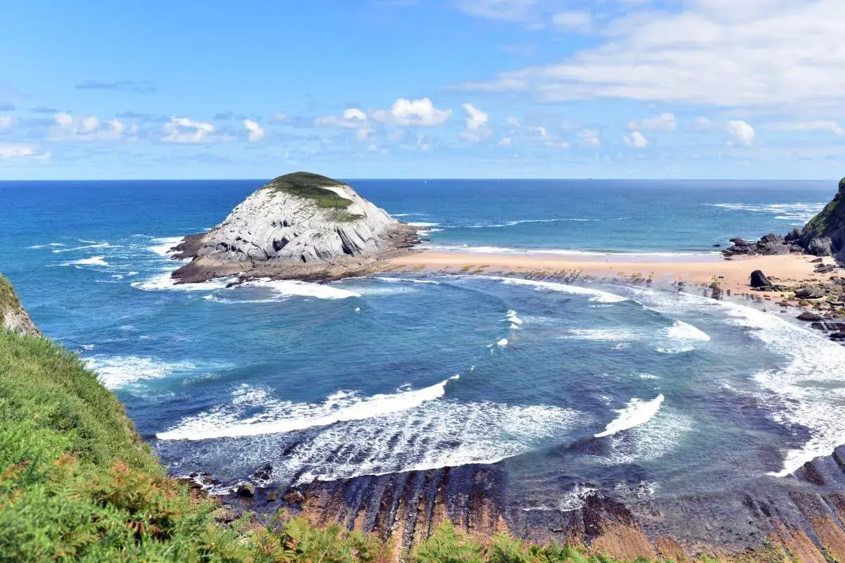 Olas rompen en las **playas limpias de Cantabria**, con un islote rocoso y vegetación verde.