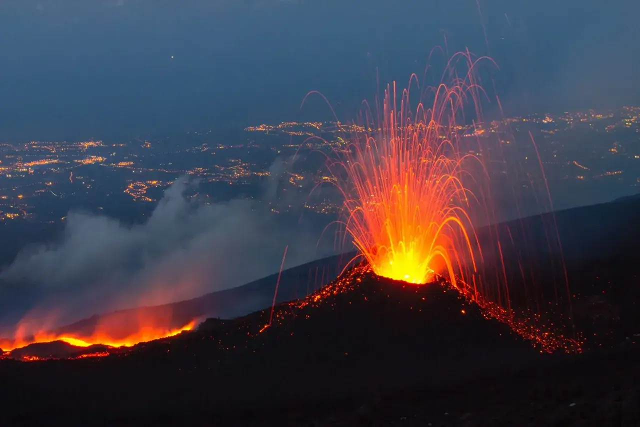 Spektakularna erupcja Etny nocą. Wulkan wyrzuca strumienie lawy i iskier na tle rozświetlonego miasta.