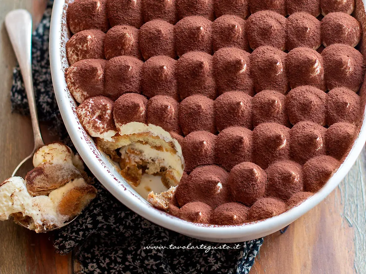 Un invitante tiramisù in una teglia bianca, cosparso di cacao. Una porzione è già stata servita su un cucchiaio, mostrando gli strati cremosi.