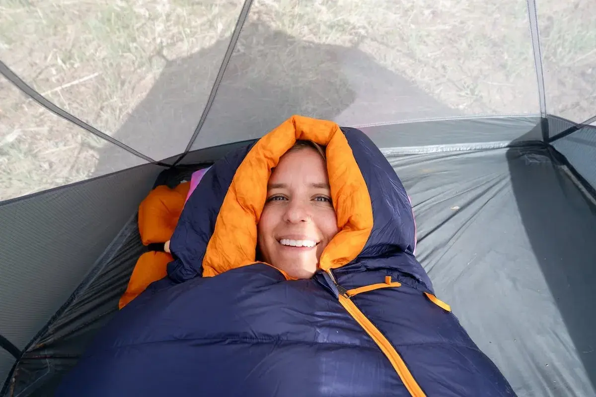 Smiling woman bundled in a navy and orange rei magma 15 weight sleeping bag inside a tent.