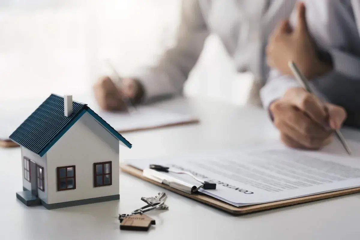 Maquette de maison, clés et contrat sur une table. Des mains signent des documents, symbolisant le **délai achat maison**.