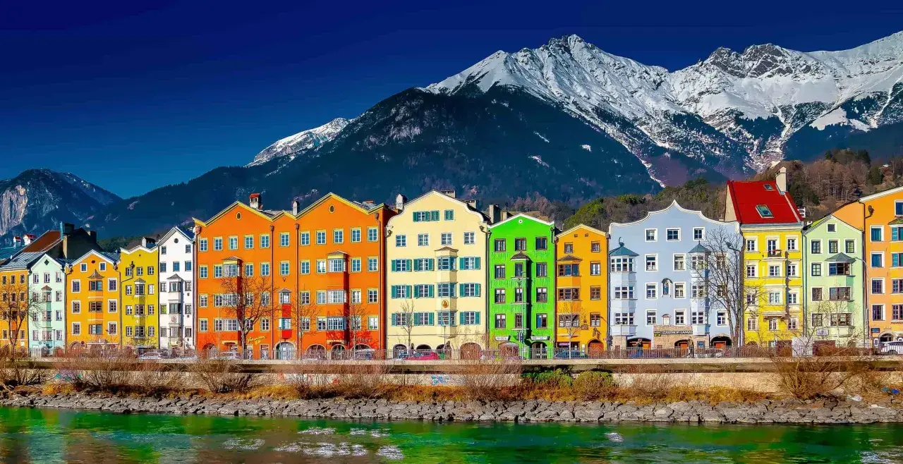 Colorful buildings line the Inn River, a vibrant scene on the journey from St. Moritz to Innsbruck, with snow-capped mountains in the background.