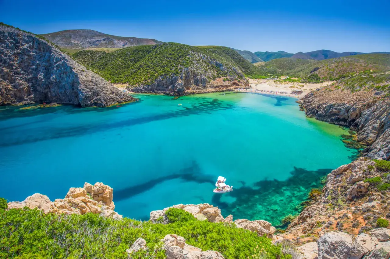 Traumhafte Cala Domestica mit türkisfarbenem Wasser, Felsen und grünen Hügeln. Ein Boot ankert in der Bucht.