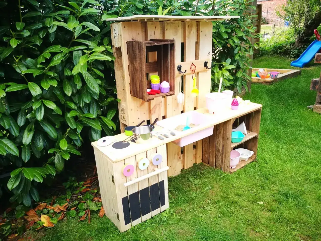 Kreative alte Weinkisten Deko Ideen: Kinderküche aus Paletten und Kisten im Garten, mit Spielzeuggeschirr und Sandkasten im Hintergrund.