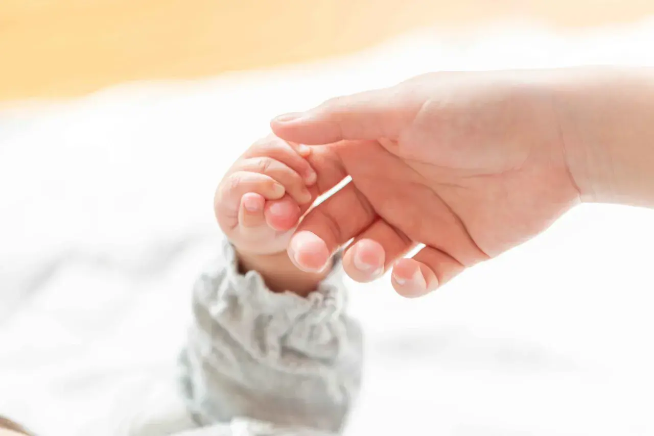 Dłoń dorosłego delikatnie obejmuje maleńką dłoń dziecka, ćwicząc chwyt pęsetkowy.