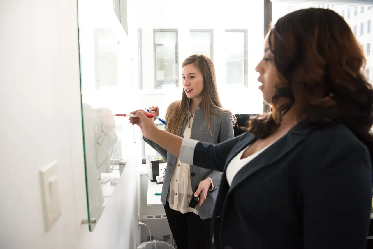 Two women discuss strategy at a whiteboard, one pointing with a marker. This scene illustrates how to ask for a promotion by preparing your case.