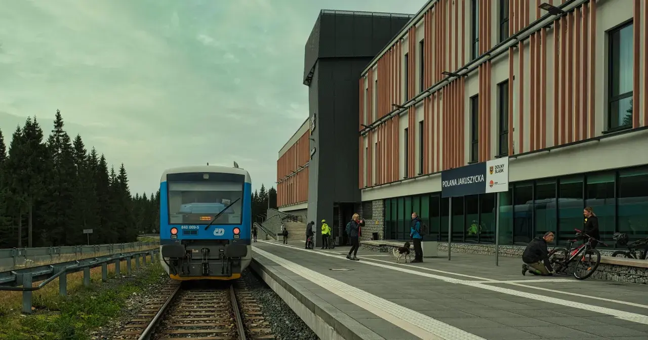 Niebieski pociąg na peronie Polany Jakuszyckiej, najwyżej położonej stacji kolejowej w Polsce. W tle nowoczesny budynek i las.