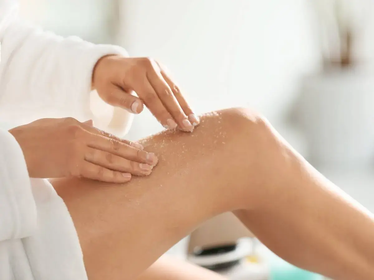 Mani che applicano scrub corpo sulla gamba, mostrando come si usa per una pelle liscia e luminosa.