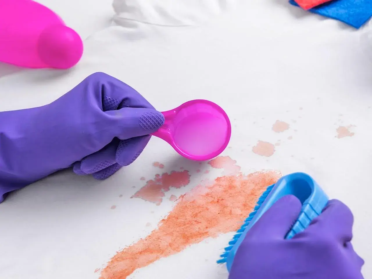 Lila Handschuhe entfernen mit Bürste und Löffel rote Flecken, vielleicht Cranberry-Flecken, von weißem Stoff.