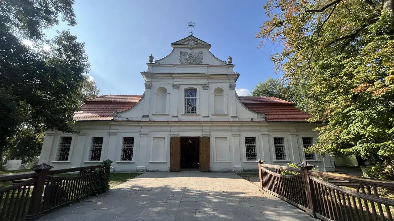 Biały kościół na wodzie w Zwierzyńcu, otoczony drzewami, z drewnianym mostkiem prowadzącym do wejścia.