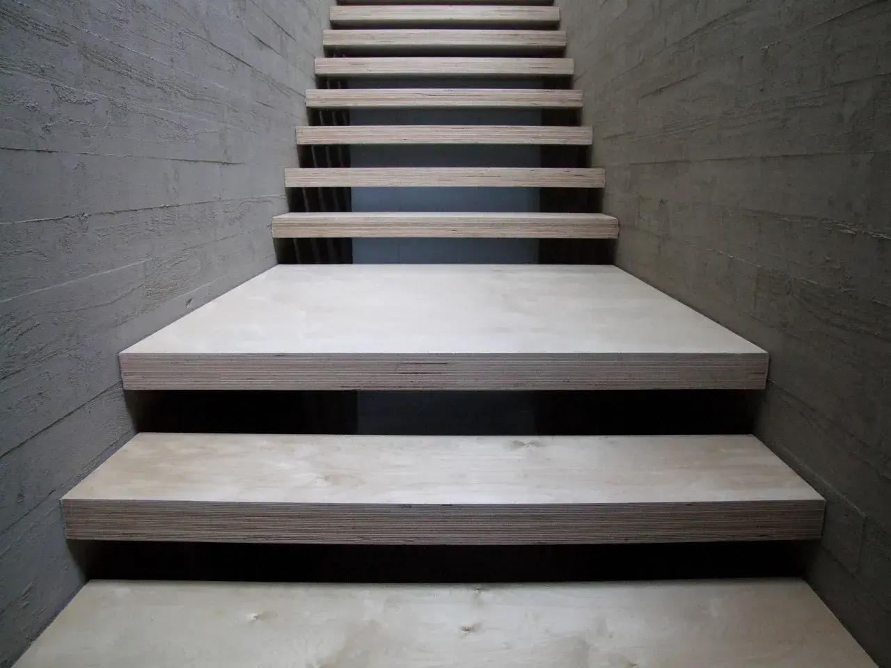 Una scala moderna con gradini in legno compensato, materiale resistente e versatile, fiancheggiata da pareti in cemento grezzo.