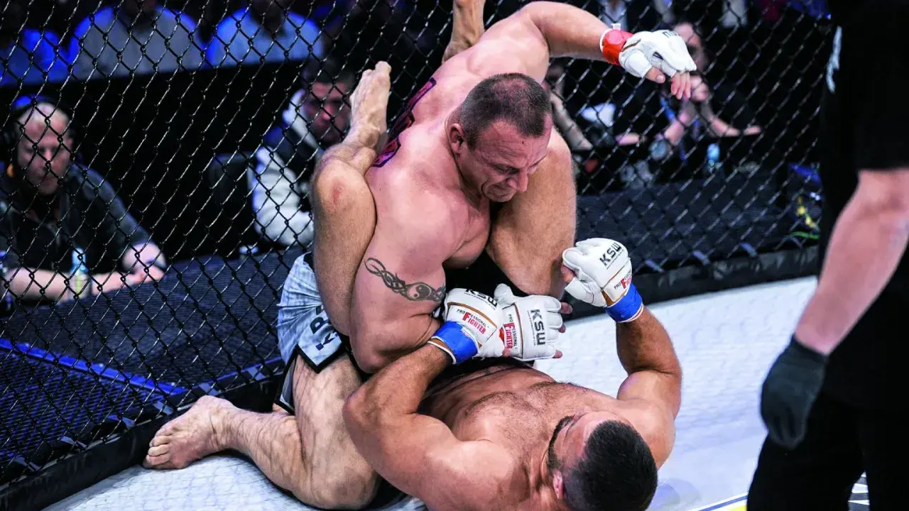 Intensywna walka Pudziana w klatce MMA. Jeden zawodnik klęczy, drugi próbuje go obalić.