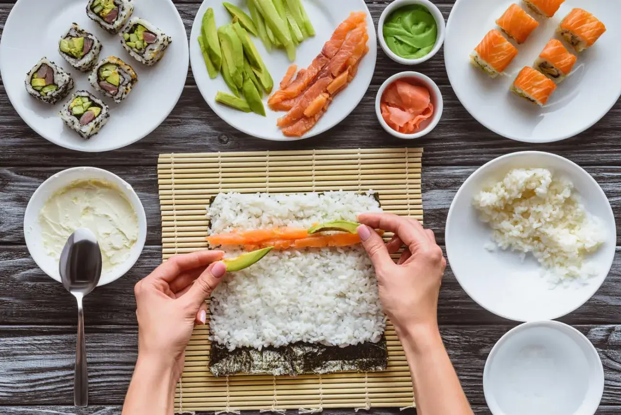 Händer lägger lax och avokado på sushiris för ett sushirecept. Ingredienser som gurka, wasabi och färdig sushi syns också.