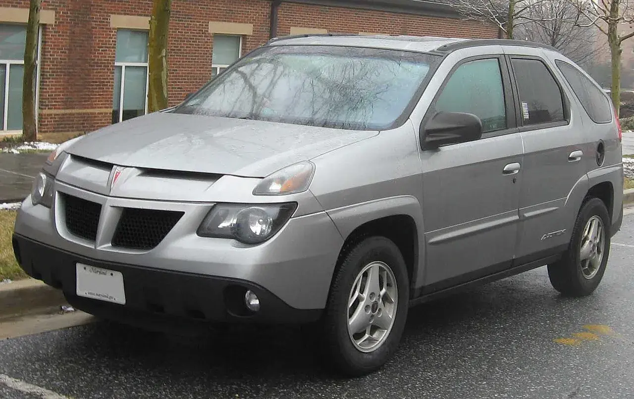 Srebrny Pontiac Aztek zaparkowany na mokrym asfalcie, z ceglanym budynkiem w tle.