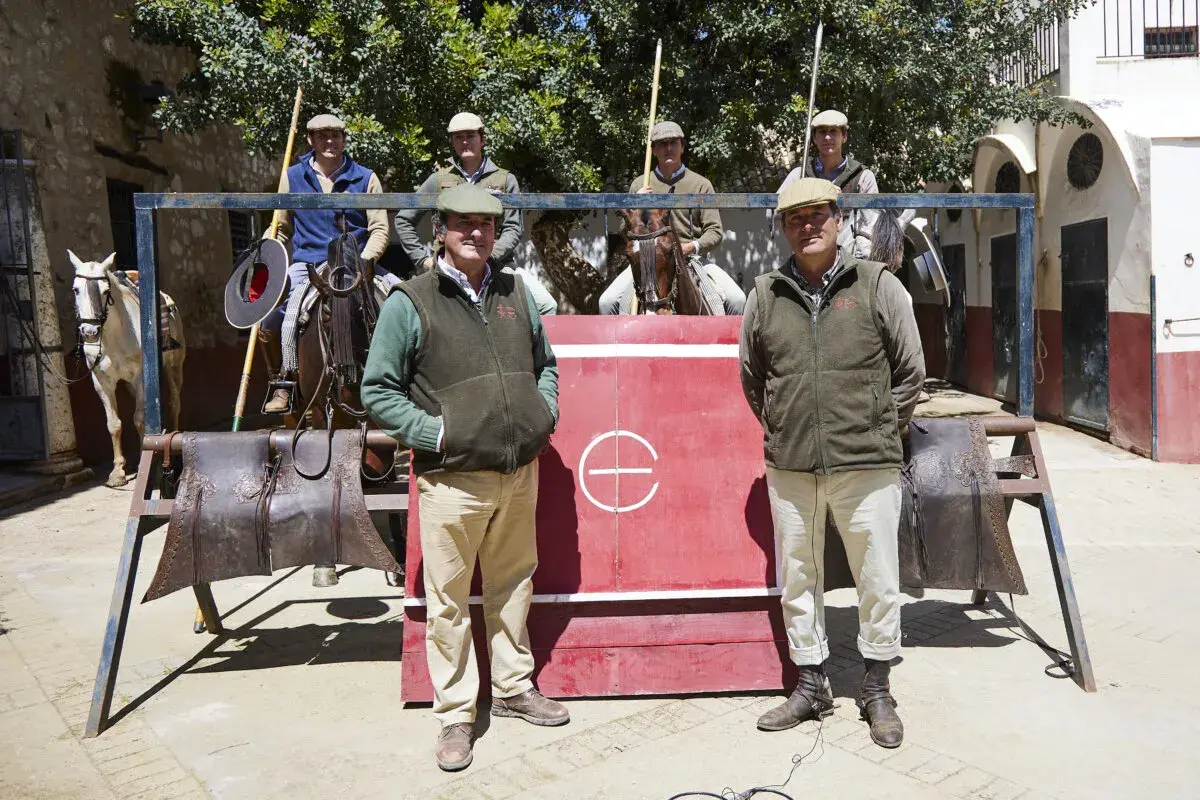 Hombres con gorras y chalecos posan frente a una barrera roja. Uno de ellos, Julio de la Puerta, sonríe. Caballos y equipo de equitación completan la escena.