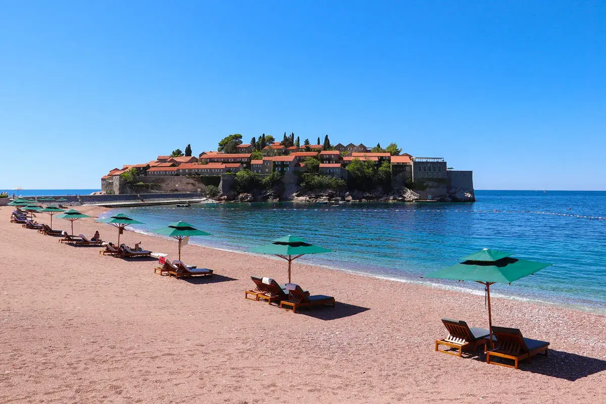 Sonnige Bucht mit rotem Sandstrand und Liegen am Meer in Montenegro. Im Hintergrund die historische Insel Sveti Stefan.