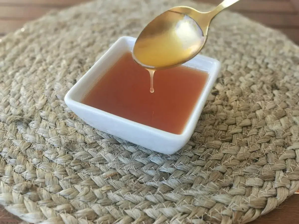 Una gota de salsa agridulce cae de una cuchara dorada a un cuenco blanco sobre una estera tejida.